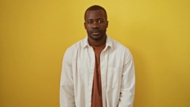 Afrika kökenli Amerikalı bir adam, sarı bir arka planda duruyor, gömlek giyiyor ve olumsuz bir jest olarak baş parmaklarını gösteriyor, mutsuz, kızgın bir yüz ifadesi var, izole edilmiş ve kötü görünüyor.