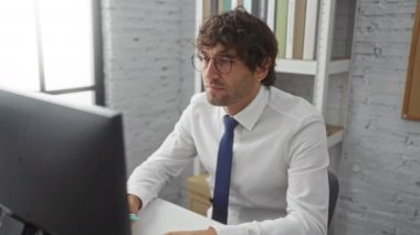 Modern bir ofiste bilgisayar başında çalışan, gözlük takan ve kravat takan, profesyonel bir iş ortamındaki görevlere odaklanmış genç bir adam..