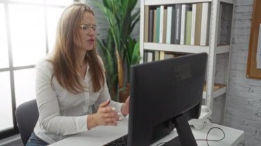 Ofisteki kadın, geniş pencereli ve kitaplıklı modern çalışma alanında otururken bilgisayardaki hayal kırıklığını dile getiriyor