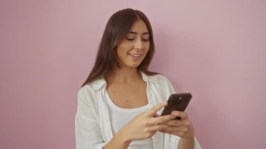 Esmer bir kadın pembe bir duvar arkasında akıllı telefonuna gülümsüyor. Neşeli bir ifadeyle ve günlük beyaz kıyafetiyle..