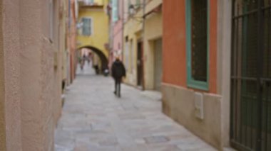 Villefranche-sur-mer 'deki bulanık yürüyüş yolu renkli binalar ve kavisli bir geçitle süslenmiş, hareketli dar bir sokakta gezinen odaksız bir figür içeriyor..