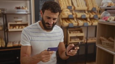 Fırındaki genç adam elinde akıllı telefon ve kredi kartıyla arka planda raflarda çeşitli fırınlanmış malların sergilendiği telefonuna bakıyor.