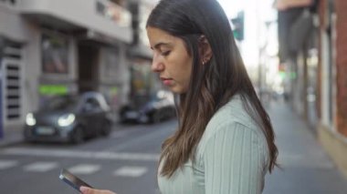Arka planda arabaları olan akıllı telefon kullanan bir kadın, gelişigüzel giyinen ve esmer saçlı genç bir kadına odaklanmış bir şehir manzarası sergiliyor..