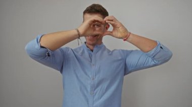 Genç adam, izole edilmiş beyaz arka plan üzerinde el kalbi oluşturuyor. Neşeli bir gülümsemeyle sevgisini ve pozitifliğini ifade ediyor. Sıradan mavi gömlek giyiyor, doğal ışıklandırma özelliklerini geliştiriyor..