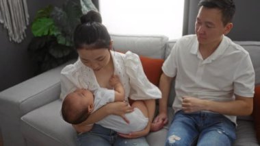 Anne ve baba rahat bir oturma odasında kanepede oturuyor, sevgi dolu bir şekilde uyuyan bebeklerini kucaklıyor, aile bağlarını ve sıcaklığını sergiliyorlar..