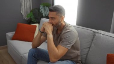 Rahat bir oturma odasındaki kanepede oturan genç adam, yumuşak yastıklarla, sıcak ışıklarla, rahat bir ortamla, tefekkür ve ev rahatlığıyla, derin düşüncelere dalarak belirir..