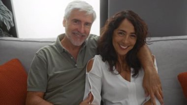 Orta yaşlı bir çift rahat bir oturma odasında birlikte oturuyor, rahat bir yuva ortamında sevgi ve mutluluğu ifade ediyorlar, kadın sıcak bir şekilde gülümsüyor..