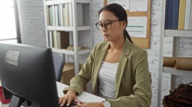 Ofisteki kadın gözlüğünü çıkarıp başını ovuyor, masa başında çalışırken baş ağrısı çekiyor, yeşil ceket ve beyaz gömlek giyiyor, etrafı kitaplar ve bilgisayarlarla çevrili..