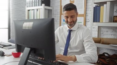 Ofisteki gülümseyen adam iş ortamında bilgisayar başında otururken akıllı telefondan video görüşmesi yapıyor..