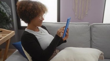Oturma odasındaki bir kanepede tablet kullanan, rahat bir ev ortamı sergileyen bir kadın..