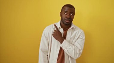 Afro-Amerikalı bir adam ayakta duruyor ve endişeli, gergin bir ifadeyle işaret ediyor, işaret parmağı bir yana, izole edilmiş sarı bir arka plan üzerinde, şaşırmış ve endişeli görünüyor