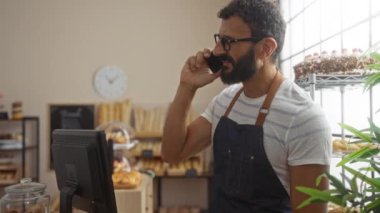 Fırında sakallı ve gözlüklü genç adam ekmek ve hamur işlerinin yanında önlükle telefonda konuşuyor.