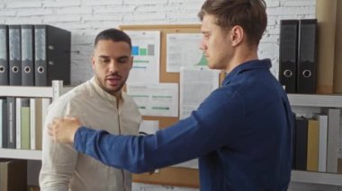 Dosyalarla dolu bir rafın yanında kahve fincanları ve dosya ve belgelerle dolu bir ilan tahtası olan iki adam tartışıyorlar.