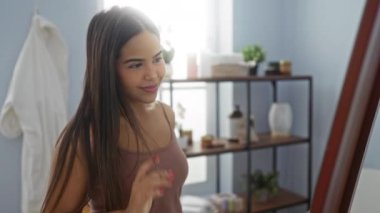 Kaplıcadaki kadın aynanın önünde saçlarını düzeltiyor. Arka planda raflar var. Kapalı alanda güzellik ve sağIık ortamı sergiliyor..