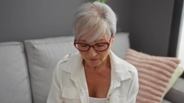 Kapalı mekanlarda gözlük tutan bir kadın modern bir oturma odasında şık dekorlu ve hafif ışıklandırmalı yorgunluk ve rahatlama belirtileri göstererek gözlerini ovalıyor.