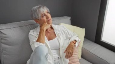 Kısa gri saçlı kadın, rahat bir oturma odasındaki gri koltukta düşünceli bir şekilde oturuyor.