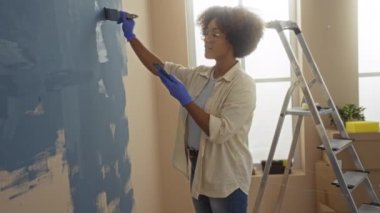 Yeni evinde duvarları mavi renklerle boyayan bir kadın, parlak bir oturma odasında merdivenin yanında eldivenler giyiyor, yaratıcılığını ve ev gelişimini gösteriyor..