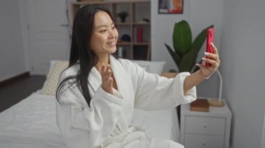 Yatak odasında, üzerinde bornozla, elinde akıllı telefonuyla, kapalı alanda gülümseyen bir kadın videosu.
