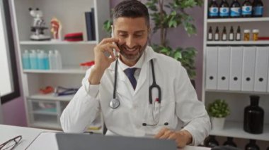 Beyaz önlüklü doktor telefonda konuşuyor. Arkaplanda malzeme rafları ve bitkiler var. Klinik masasında dizüstü bilgisayarla çalışırken gülümsüyor..