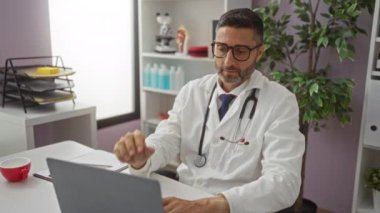 Doktor yorgunluk ve profesyonellik hissini iletirken bir klinik odasında dizüstü bilgisayar, steteskop ve bitkilerle çevrili bardakları çıkarırken yorgun görünüyor..