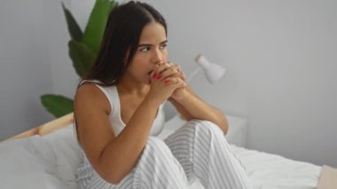 Minimalist bir yatak odasında beyaz duvarları ve çizgili çarşafları olan, stresi ve tefekkürü yansıtan düşünceli bir kadın..