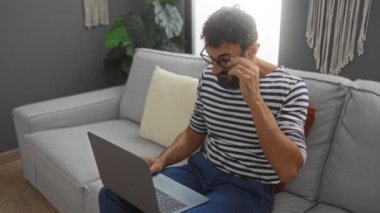 Oturma odasındaki kanepede dizüstü bilgisayarla oturan, çizgili gömlek giyen, gözlüğünü tutarken gözlerini ovuşturan, evde çalışmaktan yorulduğunu gösteren genç bir adam.