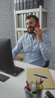 Ofiste bilgisayar başında sakallı genç adam kırmızı telefonla konuşuyor.