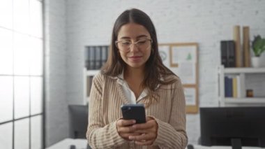 Ofis ortamında, kitaplıkları ve geniş pencereleri olan telefonu kullanarak gülümseyen bir kadın modern ve profesyonel bir çevreye işaret ediyor..