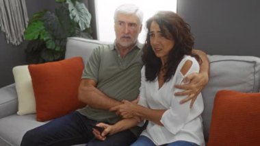 Orta yaşlı bir çift, şık bir oturma odasında rahat bir koltukta oturuyor. Birlikte televizyon izlerken, sıcak dekorlar ve kapalı hava bitkileriyle çevrili bir şekilde kaygılarını dile getiriyorlar..