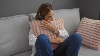Esmer saçlı, oturma odasında oturan, kulaklık takan, saat ve kot pantolon giyerken pembe yastığına sarılan genç bir kadın.