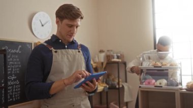 Bir fırında çalışan erkekler dijital tablet ve pastanede sergilenen pastalarla, kapalı bir dükkanda ekmek menüsü ve arka planda bir saatle