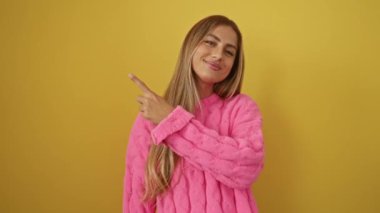 Süveter giyen genç sarışın kadın, neşeli bir şekilde gülümserken izole edilmiş sarı arka planı işaret ediyor.