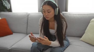 Kulaklık takan bir kadın evdeki koltukta oturmuş modern bir oturma odasında telefonuyla oyun oynuyor.