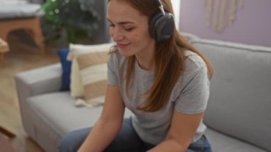 Kadın, rahat oturma odasında kulaklık takarak kanepede dinleniyor ve evde gündelik müzik dinliyor.