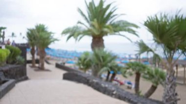 Lanzarote 'ta bulanık bir açık hava sahnesi. Arka planda palmiye ağaçları, patikalar ve dağılmış insanlarla dolu bir turist plajı..