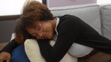 Kadın rahat bir oturma odasında rahat rahat, kıvırcık saçlı, gözlüklü, yastığa yaslanmış ev ortamında huzurlu bir şekilde gülümsüyor..