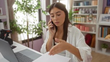 Ev dekorasyonu dükkanındaki çekici esmer kadın masadaki belgelere bakarken telefonla konuşuyor.