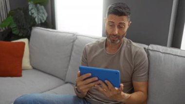 Adam, modern oturma odasında tabletle kanepede dinleniyor, odaklanmış ve rahatlamış, rahat yastıklar ve kapalı bitkilerle çevrili, memnuniyet ve ilgi ifade eden.