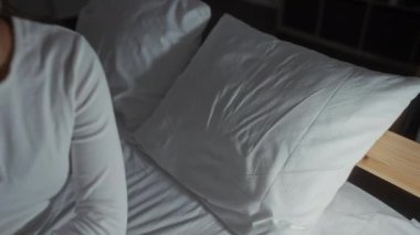 Genç bir kadın, esmer saçlı, beyaz tişörtlü, evinde dinleniyor ve rahat bir yatak odasında, yumuşak bir ışık altında yakılıyor..
