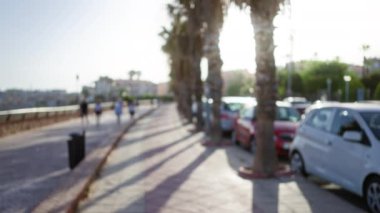 Parlak güneşli bir günde, kaldırımda palmiye ağaçları ve park etmiş arabalarla yürüyen insanların odağını kesmiş.
