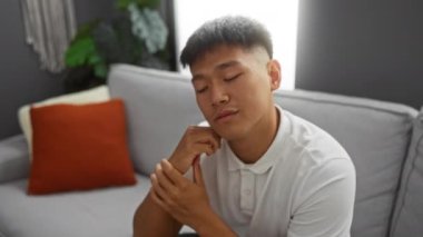 Modern bir oturma odasındaki kanepede dinlenen genç adam beyaz gömlekli ve rahat bir ev ortamında kanepede turuncu yastıklı.