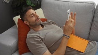 Evdeki koltukta oturan, elinde telefon ve turuncu kitapla modern bir apartman dairesinde boş vakitlerin tadını çıkaran bir adam..