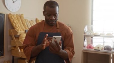 Fırının içinde kapalı yumruklarla kutlayan bir adam, akıllı telefon kullanırken gülümseyen, saatin altında ekmek ve hamur işleriyle çevrili bir adam..