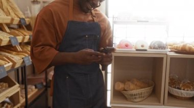 Fırında çalışan genç adam gülümseyen bir telefon kullanıyor. Etrafı ekmek ve pastalarla çevrili. İçerideki rahat atmosferi gösteriyor.