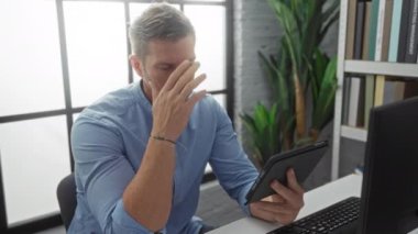 Ofisteki genç adam elinde tablet tutarken sıkıntılı görünüyor; işyeri sahnesi, bitki ve kitaplarla modern içimdeki stresi resmediyor, erkek profesyonel zorlukları vurguluyor.