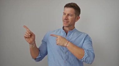 Mavi gömlekli genç adam özgüvenle parmaklarını beyaz bir arka plana doğru uzatıyor. Stüdyo ortamında kendine güveni ve cazibesi var..