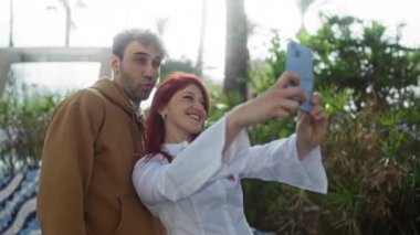 Açık havada, parlak, güneşli bir günde yeşil yeşilliklerin ve kentsel geçmişin ortasında akıllı telefonlu neşeli bir selfie çekiyorlar..