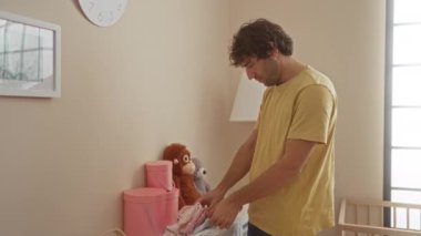 Genç bir adam rahat bir yatak odasında bebek kıyafetlerini katlıyor, hafif ışıklandırmayla, bir yuva ortamında bebek için hassas bir hazırlık ve bakım anı gösteriyor..