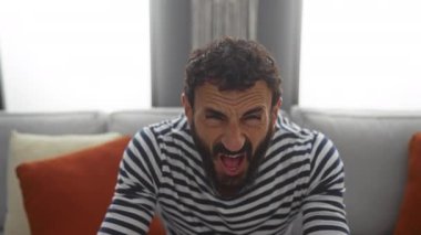 Çizgili gömlekli sakallı, hayal kırıklığına uğramış bir adam oturma odasında kanepede oturuyor.