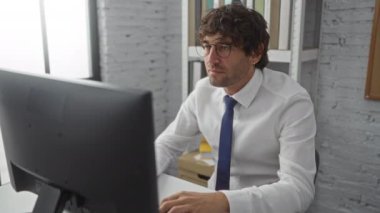 Ofisteki genç adam gözlük ve kravat takıyor, elleri başının üzerinde stresli görünüyor, bilgisayarla masada oturuyor, arka planda kitaplık var, işindeki hayal kırıklığını gösteriyor..